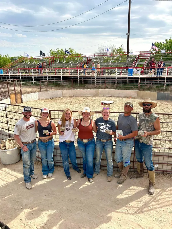 Seven Ham Rodeo buckle winners in the arena ring, flags flying over the bleachers behind them.