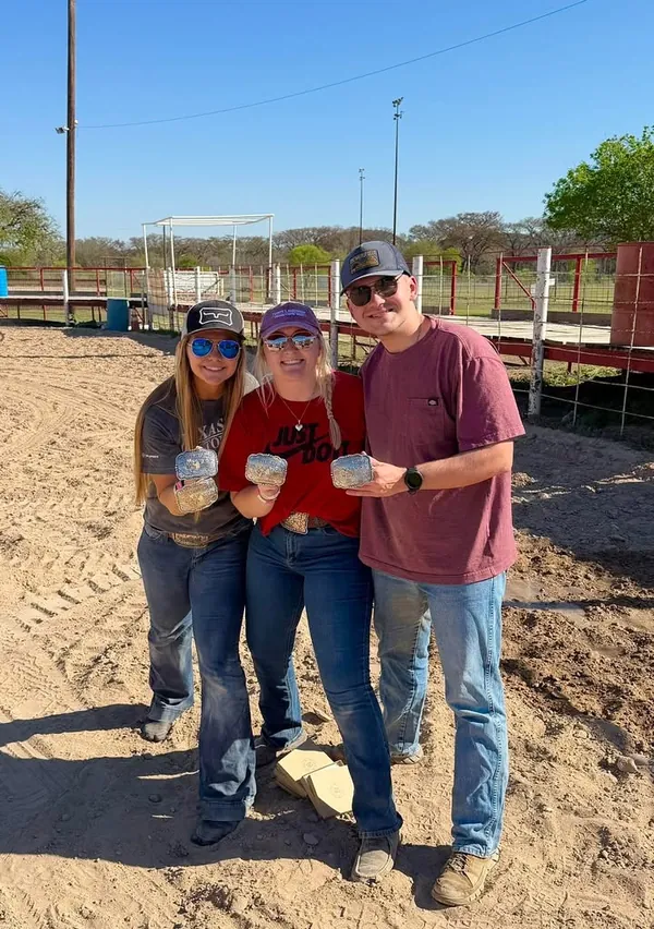 Three smiling Ham Rodeo winners hold up their buckles under a blue sky.