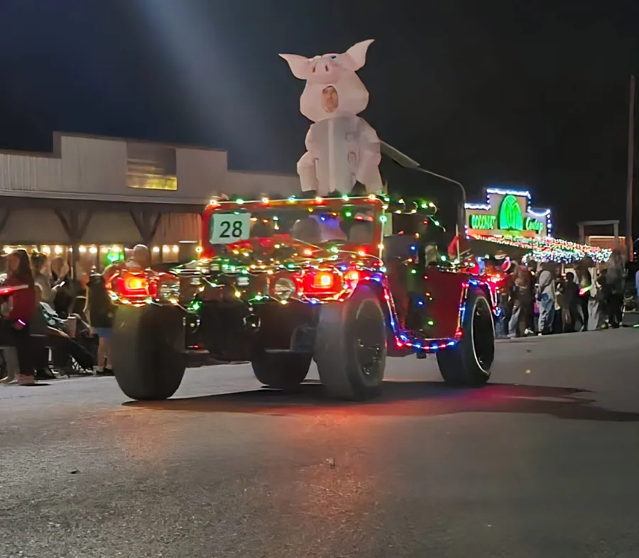 A truck strung with Christmas lights, with a pig mascot riding on top, in the Bandera Christmas parade at night.